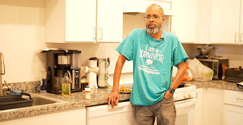 A man stands in his kitchen wearing a blue shirt that says "survivor"